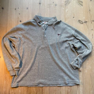 Grå pikétröja från Nike - Säljer en grå långärmad pikétröja från Nike med broderad logga på bröstet och svart Nike-text på kragen. Tröjan har klassisk krage och knappar framtill. Köptes vintage och är i högsta grad det! 
