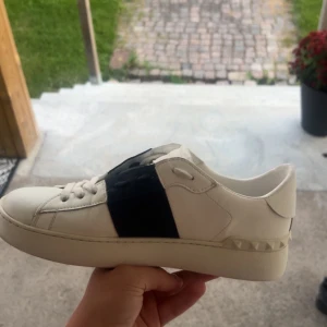 mörkblåa Valentina open - Snygga Valentino Open sneakers i vitt skinn med en bred blå rem över mitten och coola pyramidnitar på hälen. Klassisk rund tå och platt sula, perfekt för dig som gillar stilrena och lyxiga detaljer. Skorna har snörning och är riktigt bekväma. Box ingår inte. Byten funkar också.