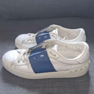 Valentino Garavani Open sneakers vit/blå - Säljer ett par Valentino Garavani Open sneakers i vitt skinn med blått band över snörningen. Snygga nitar längs hälen och platt sula. Klassisk rund tå och cool design som sticker ut. Perfekt för dig som gillar lyxiga detaljer och streetstyle. Obs lite målade!