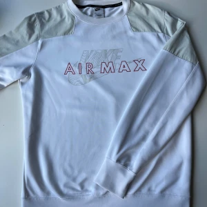 Nike Air Max vit sweatshirt - Nike Air Max sweatshirt i vitt med ljusgrå axelpartier och röd logga framtill och baktill. Tröjan har lång ärm, rund hals och tryckt Air Max-design på bröstet. Perfekt för dig som gillar sportig och clean stil. Materialet är mjukt och skönt bomullsmix.