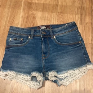 Blå jeansshorts med spets från Superdry - Snygga blå jeansshorts från Superdry med vit spetskant längst ner. Modellen är lågmidjad och har klassiska fickor och knappstängning framtill. Perfekt till sommaren och ger en chill vibe med den loose passformen och coola spetsdetaljen. Pris går absolut att diskutera!