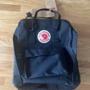 Svart Fjällräven Kånken ryggsäck - Klassisk svart Fjällräven Kånken ryggsäck med ikonisk rund logga i rött och vitt framtill. Väskan har ett stort huvudfack, en mindre ficka med knapp framtill och justerbara axelremmar. Materialet är slitstark polyester och designen är stilren och fyrkantig.