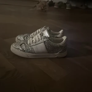 Valentino Garavani flycrew - Säljer ett par Valentino Garavani sneakers med silverglittrigt tyg och vita skinnpaneler. Skorna har rund tå, platt sula och snörning. Insidan är vit och mjuk, och yttersulan har små nitar baktill. Perfekta för dig som vill sticka ut med extra bling. Kan diskutera pris!! Ena skon är lite trasig. 