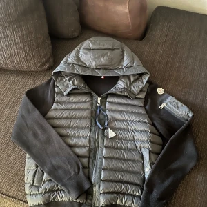 Svart cardigan med huva från Moncler - Snygg svart cardigan från Moncler med stickade svarta ärmar och vadderad kropp. Västen har huva, dragkedja framtill, fickor med tryckknapp och en liten ärmficka med dragkedja. Klassisk Moncler-logga på ärmen, perfekt till hösten!