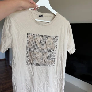 Beige t-shirt Karl Lagerfeld M - Snygg beige t-shirt från Karl Lagerfeld i storlek M. Framsidan har ett coolt grafiskt tryck med pixelmönster och text. T-shirten är kortärmad och har en klassisk rund hals. Materialet är mjuk bomull som känns skönt mot huden.