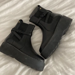 Svarta boots med äkta skinn - Säljer ett par svarta boots med rund tå och rejäl plattformsula. Skorna har snygga spänndetaljer på sidan och en diskret dragkedja för enkel påtagning. Materialet är äkta skinn och insidan har en grå textildetalj vid skaftet. Perfekt för dig som gillar chunky stil.