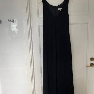 Svart långklänning från Bandolera - Elegant svart långklänning från Bandolera med v-ringad front och ärmlös design. Klänningen har en diskret slits framtill och dragkedja i ryggen för enkel påklädning. Materialet är mjukt och följsamt, perfekt för dig som gillar stilren och tidlös look. Snyggt fall med sprunn från bröstet och nedåt fram och kortare klänning under, se bild 3. Använd ett fåtal tillfällen 