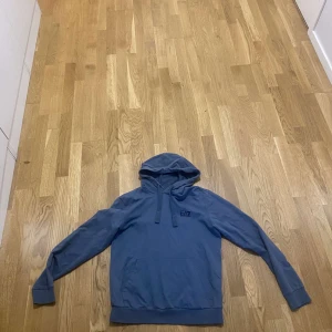Blå hoodie från EA7 med huva - Snygg blå hoodie från EA7 med huva och snörning. Tröjan har en klassisk känguruficka framtill och långa ärmar med muddar. Perfekt för chill dagar eller när du vill ha en avslappnad stil. EA7-logga på bröstet ger en sportig touch.