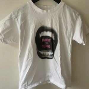 Vit t-shirt Hungry For Techno - Snygg vit t-shirt med tryck av en öppen mun och texten 'HUNGRY FOR TECHNO' i rosa. Klassisk rund hals och korta ärmar, perfekt för dig som gillar statement-plagg och elektronisk musik. Materialet känns som bomull och passformen är normal.