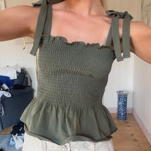 Olivgrönt linnetopp med volang - Supersöt olivgrön linnetopp med smockad överdel och volang nedtill. Axelbanden knyts med rosett och ger en chill vibe. Toppen har fyrkantig halsringning och är ärmlös, perfekt för soliga dagar. Materialet känns mjukt och lätt mot huden. Säljer även kjolen!