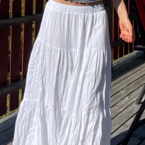 Vit långkjol i bomull - Säljer en vit långkjol med volanger och resår i midjan från stradivarius. Kjolen är i storlek xs, gjord i mjuk bomull och har en luftig, flowy känsla som passar perfekt till varma dagar. Den är helfärgad och har en snygg, avslappnad siluett. Säljer även samtliga toppar