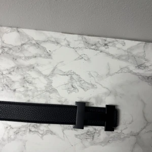 Svart bälte  - Snyggt svart bälte med ett stilrent H-format spänne i matt svart metall. Bältet är tillverkat i präglat skinn med en klassisk, rak form som ger en lyxig vibe. Perfekt för dig som vill ha en statement-accessoar med exklusiv känsla.