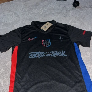 Svart FC Barcelona x Nike fotbollströja - Snygg svart FC Barcelona x Nike fotbollströja med korta ärmar och Dri-FIT material. Tröjan har röda och blå sidopaneler, klubbmärke på bröstet och coolt tryck 'Cactus Jack'. UNHCR-logga på ryggen och Nike Swoosh i rött. Perfekt för dig som gillar streetstyle och fotboll.