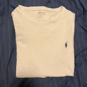 Vit t-shirt från Polo Ralph Lauren - Vit t-shirt från Polo Ralph Lauren med klassisk rund hals och korta ärmar. Diskret blå Polo-logga broderad på bröstet. T-shirten är i mjuk bomull och har en normal passform som sitter snyggt på kroppen. Perfekt för en clean och stilren look.