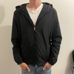 Svart vindjacka/windbreaker från Moncler med huva - Svart vindjacka från Moncler med huva. En liten defekt, skriv för bild, annars bra skick. Storlek M men passar större och mindre skulle jag säga. Inga snören då jag tyckte det var snyggare utan. Inget äkthets bevis, men NFC-Scan, och så har jag själv legitcheckat den så e ganska säker på att den är äkta.
