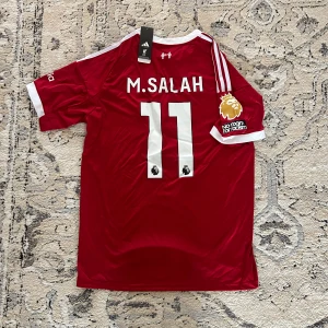 Liverpool Salah #11 röd matchtröja - Liverpool FC matchtröja med M. Salah och nummer 11 på ryggen. Röd med vita detaljer, kortärmad och tillverkad i lätt polyester. Adidas-logga, klubbmärke och Premier League patch på ärmen. Snygga vita ränder på axlarna och sponsortryck framtill.