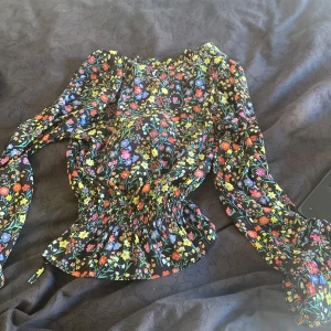 Blommig blus med puffärmar - Superfin blommig blus med svart botten och färgglada blommor i rött, gult, blått och lila. Blusen har puffiga långärmar och smockad midja som ger en snygg siluett. Perfekt för dig som gillar färg och mönster!