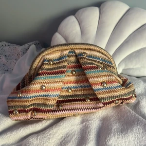 Randig kuvertväska med nitar - Säljer en unik kuvertväska med färgglada ränder i beige, rosa, blått, rött och grönt. Väskan är stickad och har coola guldfärgade nitar som detaljer. Perfekt storlek för det viktigaste och ger din outfit en personlig touch. Band ingår!