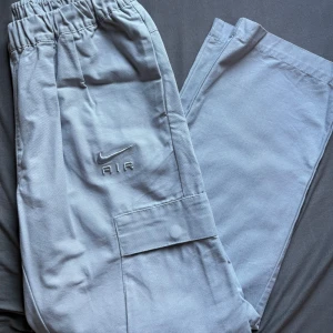 Nike Air ljusgrå cargopants S - Nike Air cargopants i ljusgrått med broderad logga på benet. Byxorna har elastisk midja, stora sidofickor och en loose passform. Tillverkade i mjuk bomull, perfekta för en chill streetwear-look. Snygga detaljer och riktigt skön vibe. Baggie och passar större storlekar, använd väldigt lite, några gånger