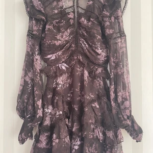 Mörklila blommig volangklänning med spets - Säljer en mörklila kortklänning med lila blommigt mönster och romantiska volanger. Klänningen har långärmade puffärmar, spetsdetaljer och omlottliknande rynk framtill. Materialet är lätt och transparent, perfekt för dig som gillar en bohemisk och feminin vibe.