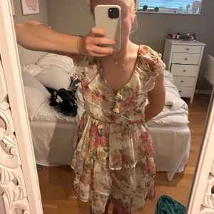 Blommig kortklänning med volanger - Superfin blommig kortklänning i ljusa färger med rosa och gröna toner. Klänningen har volangdetaljer, v-ringning och öppen rygg med en tunn rem. Från nelly! Storlek 34