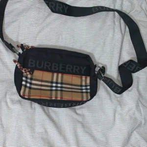 Burberry väska - Snygg axelväska från Burberry med klassiskt rutigt mönster i beige, svart, vitt och rött på framsidan. Väskan har en bred svart axelrem med Burberry-logga och detaljer i silver. Materialet är polyester och väskan har dragkedja upptill.