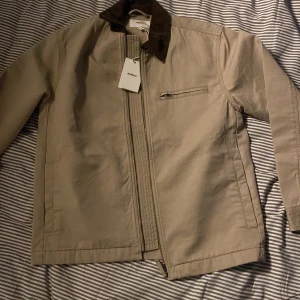 Beige overshirt från Dobber - Snygg beige overshirt från Dobber med mörkbrun manchesterkrage och diskret bröstficka med dragkedja. Jackan har raka linjer, lång ärm och stängs med dragkedja framtill. Perfekt för dig som gillar stilrena och klassiska plagg.
