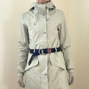 TRETORN REGNKAPPA/ TRENCHCOAT - TVÅ KAPPOR I EN- Lika användbar som trenchcoat soliga dagar som regnkappa när det öser ner. Knappt använd alls, lätt och följsam, 100 % vatten- & vindsäker. Perfekt huva, två stora fickor fram, Medföljer utan kostnad: Ralph Lauren-skärp som jag låtit sy fast i två hällor bak så skärpet inte glider ur när man hänger upp kappan. Originalskärp medföljer. Inga fläckar, helt felfri, storlek S. . Kostat ca 1200.