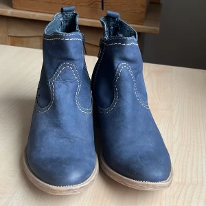 Blå boots från Tamaris - Snygga blå boots från Tamaris med dekorativa sömmar och perforerade detaljer på sidorna. Skorna har en bekväm klack och dragkedja för enkel påtagning. Perfekta för en stilren look.