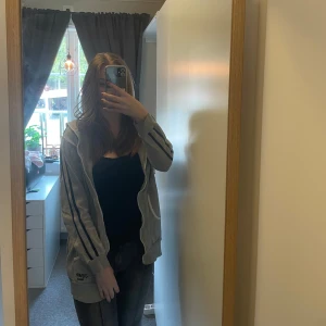 Grå huvtröja från Nike - Säljer en grå huvtröja från Nike med dragkedja och svarta ränder längs ärmarna. Tröjan har en luva och fickor framtill, perfekt för en avslappnad stil. På ryggen står det ”UNITED 1966”.