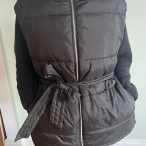 Svart dunväst med knytband - Snygg svart dunväst med dragkedja och knytband i midjan. Perfekt för lager-på-lager under kyliga dagar. Västen har en stilren design och är lätt att matcha med olika outfits.