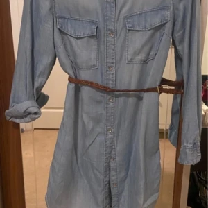  jeansklänning  - Säljer en stilren ljusblå jeansklänning hm . Klänningen har långa ärmar och knappar framtill samt två bröstfickor. Den kommer med ett flätat bälte som ger en snygg passform. Perfekt för en avslappnad look!
