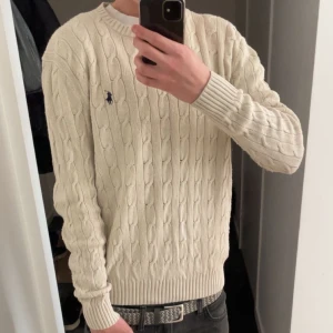 Kabelstickad från Ralph Lauren - Säljer en stilren vit stickad tröja från Polo Ralph Lauren med kabelstickat mönster. Tröjan har långa ärmar och en broderad logotyp på bröstet. Perfekt för en klassisk look. Storlek: S, Skick: 10/10