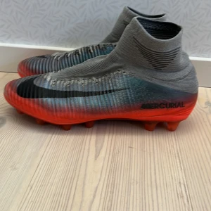Nike Mercurial fotbollsskor - Snygga Nike Mercurial fotbollsskor i grått och rött med hög ankelkrage för extra stöd. Det är i bra skick förutom att det saknar snören och en liten slitning på framsidan av skon med det är inget som håller på och gå sönder