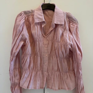 Rosa blus från Dazy - Säljer en söt rosa blus från DAZY med långa ärmar och knappar framtill. Blusen har en rynkad design som ger en fin struktur och volym. Perfekt för en stilren look.