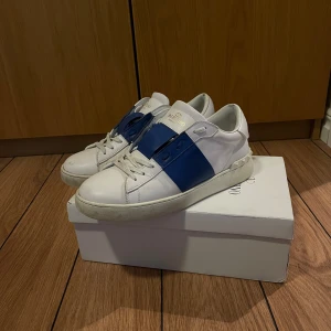 Valentino skor - Snygga Valentino Garavani sneakers i vitt med en iögonfallande blå rem över snörningen. Perfekta för att ge din outfit en lyxig touch. Skorna kommer med originalförpackning och dustbag.