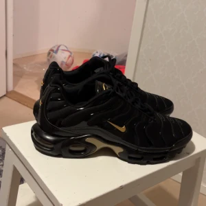 Nike Tn skor  - Snygga svarta Nike sneakers med gulddetaljer och Tn-logga på hälen. Skorna har en sportig design med synlig dämpning i sulan och en elegant svart finish. Perfekta för en stilren look.