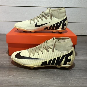 Beige Nike Mercurial fotbollsskor - Säljer ett par beige Nike Mercurial fotbollsskor med svart logga och snörning. Skorna har en rund tå och är designade för optimal prestanda på planen. Perfekta för fotbollsentusiaster som vill ha stil och funktionalitet.