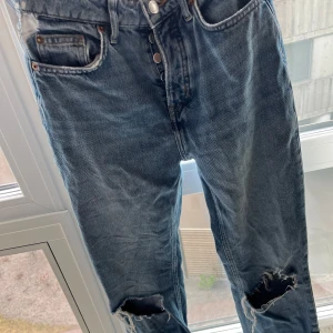 Blå jeans med slitningar - Säljer ett par blå jeans med coola slitningar på knäna. De har en klassisk femficksdesign och knappgylf. Perfekta för en avslappnad stil.