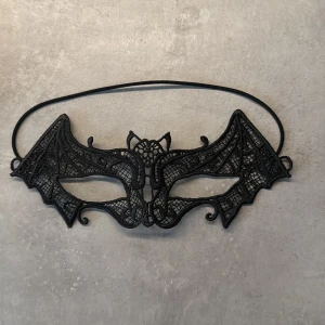Svart spetsmask med fladdermusdesign - Elegant svart mask i spets med fladdermusdesign, perfekt för maskerad eller fest. Masken har detaljerade spetsmönster och hålls på plats med ett elastiskt band.