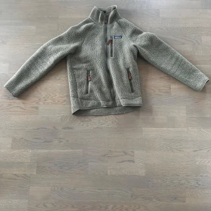 Fleecejacka från Patagonia - Säljer en beige fleecejacka från Patagonia med dragkedja och hög krage. Jackan har två framfickor med dragkedjor och en liten bröstficka. Perfekt för kyliga dagar och outdoor-äventyr.