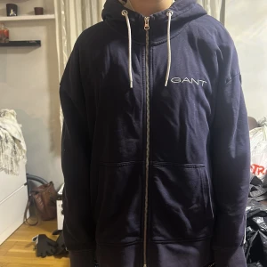 Mörkblå hoodie från GANT - Säljer en snygg mörkblå hoodie från GANT med dragkedja och justerbar huva. Den har ett stilrent tryck med GANT-loggan på bröstet och ryggen. Perfekt för en avslappnad stil.