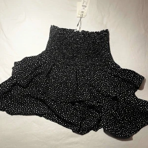 Svart prickig kjol - Säljer denna fina svarta kjol med vita prickar och volangdetaljer. Prislapp finns kvar.