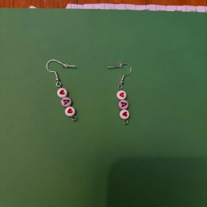 Örhängen med bokstäver och tärningar - Två par unika örhängen. Ett par med bokstäver och röda hjärtan på vita pärlor. Det andra paret har orange tärningar och små korsdetaljer i guld.