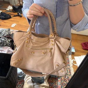 Ljusrosa balenciaga väska (intressekoll) - Säljer min älskade ljusrosa balenciaga med guldfärgade nitar och dragkedja. Väskan har både handtag och en avtagbar axelrem för flexibilitet. Perfekt för att bära dina dagliga nödvändigheter med stil.