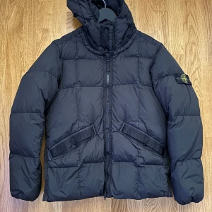 Svart dunjacka från Stone Island - Snygg svart dunjacka från Stone Island med huva och dragkedja framtill. Jackan har en quiltad design och två fickor med knappar. Perfekt för kyliga dagar.