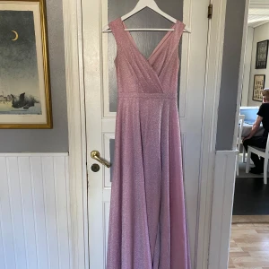 Rosa glittrig balklänning - Elegant rosa balklänning med glittrigt tyg och omlott-design. Klänningen är ärmlös och har en v-ringad framdel, perfekt för festliga tillfällen såsom balen. Lite smutsfläckar innanför klänningen 