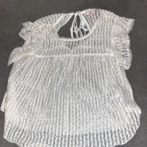 Spetsblus/knytblus - Säljer en söt vit blus från Nelly med spetsdetaljer och volanger. Blusen har korta ärmar och en knytning i ryggen. Perfekt för en somrig look!