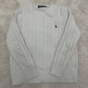 Vit stickad tröja  - Säljer en stilren vit stickad tröja  med kabelstickat mönster och broderad logga på bröstet. Tröjan har långa ärmar och ribbade muddar vid ärmslut och nederkant. Perfekt för kyliga dagar! Storlek M/S typ.