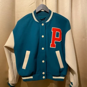 Blå och vit bomberjacka från Gina Tricot - Snygg blå och vit bomberjacka från Gina Tricot med röd bokstav 'P' på bröstet och 'PARIS' på ryggen. Jackan har vita ärmar och stängs med knappar. Perfekt för en cool och avslappnad stil.